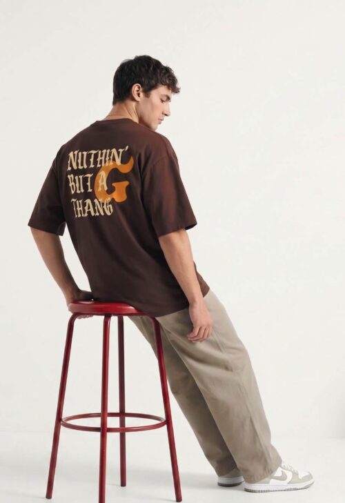 Man met bruine oversized T-shirt met print en beige broek, zijwaarts zittend op rode kruk tegen witte achtergrond