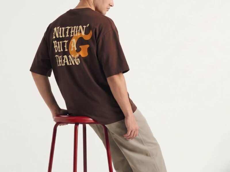 Man met bruine oversized T-shirt met print en beige broek, zijwaarts zittend op rode kruk tegen witte achtergrond