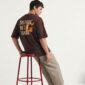 Man met bruine oversized T-shirt met print en beige broek, zijwaarts zittend op rode kruk tegen witte achtergrond