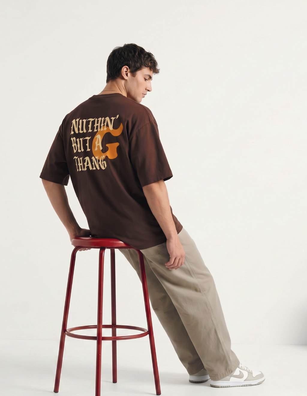 Man met bruine oversized T-shirt met print en beige broek, zijwaarts zittend op rode kruk tegen witte achtergrond